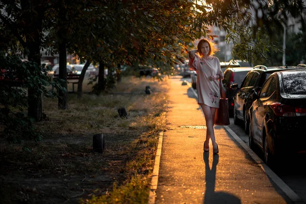 一位40岁的成年妇女穿着晚霞中的连衣裙沿街走着 路上停着汽车 — 图库照片