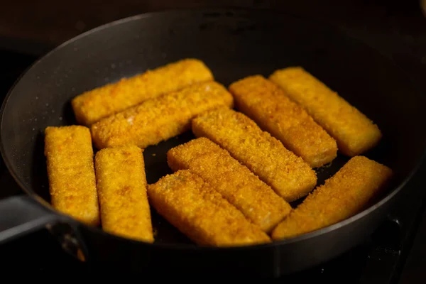Breaded fish sticks in a frying pan. Preparation of frozen fish sticks. Fast food. Dark background.