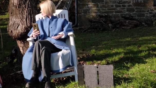 一个女人悲伤地坐在户外拿着一部手机 附近是绿色草地上的便携式太阳能电池 凉爽的晴天 — 图库视频影像