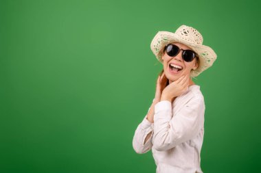 Yeşil arka planda beyaz gömlekli, şapkalı ve güneş gözlüklü neşeli bir kadın. Metnin için yer.