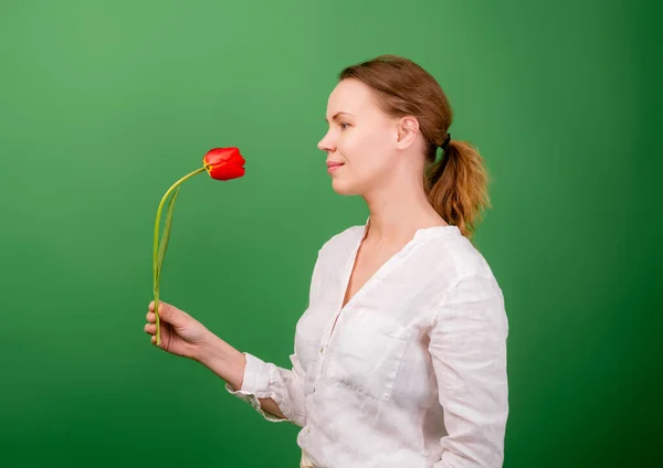 stock image A pretty woman of middle age forty years old holds one flower - a red tulip on a green background.