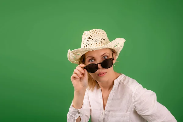 戴着帽子 戴着太阳镜 身穿绿色背景白衬衫的快乐而惊讶的女人 看着摄像机 把眼镜放下 — 图库照片