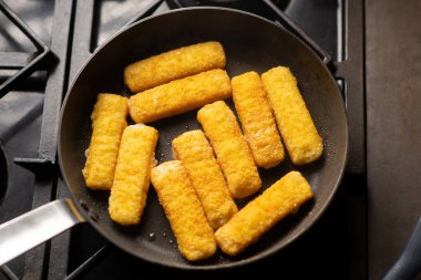 Breaded fish sticks in a frying pan. Preparation of frozen fish sticks. Fast food. Dark background.