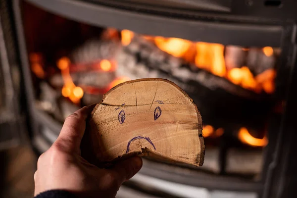 Rise in price of firewood for heating concept. Hand holds log with sad smiley against background of burning fireplace