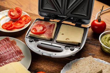 Sandviç makinesinde sandviç pişirmek. Farklı malzemeler. Ekmek, peynir, ahşap arka planda sebze..