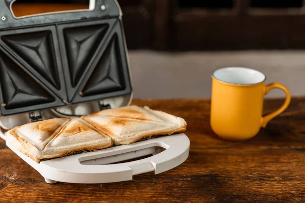 Tahta bir arka planda sandviç makinesinde yeni yapılmış tost sandviçler. Yanında bir fincan daha var. Sabah kahvaltısı kavramı.