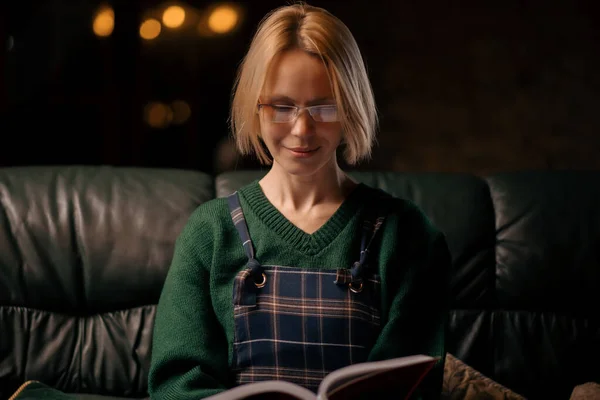眼鏡をかけた女性が家のソファの上で本を読んでいる 40歳の女性は魅力的な本を読んでいます — ストック写真