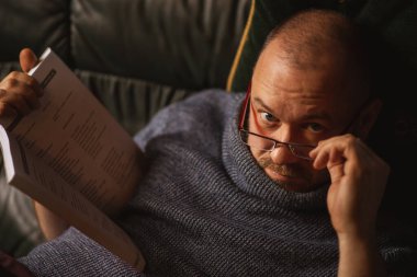 Gözlüklü bir adam kanepede yatarken kitap okuyor. Sıradan görünümlü orta yaşlı bir adam evde günlük giysiler içinde. Okuma, öğrenme.