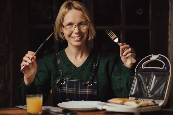 女は微笑んで朝食を待つ 彼女はナイフとフォークを手に持っている サンドイッチを作る 食べる — ストック写真