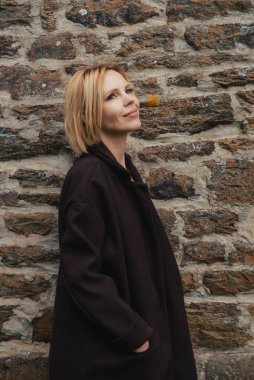 Taş bir duvarın arkasında ceketli bir kadın. Kırk yaşında orta yaşlı bir kadın. İnce yapılı..