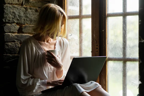 A woman sits with a laptop near the window. A middle-aged woman communicates, works at a laptop. Freelancer, work from home.