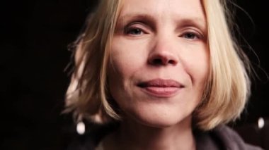 Orta yaşlı bir kadın gülümsüyor ve siyah arka planda kameraya yakından bakıyor..