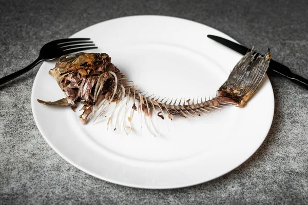 stock image Fried fish skeleton, spine and bones on a white plate with a black knife and fork on a gray background.