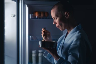 Kısa saçlı bir kadın geceleri buzdolabının yanında yemek yer. Gece iştahı, diyet konsepti. Kel, zayıf bir kadın açlığını gideriyor..