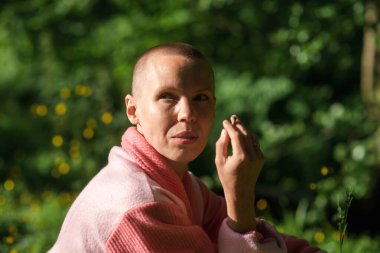 Bornozlu kel bir kadın yeşil bitki arka planında sigara içiyor. Güneşli bir gün.