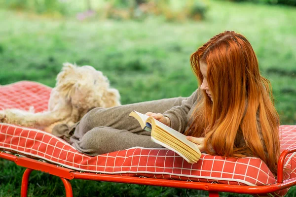 A young girl with a dog reads a book lying on the outdoors bed. Rest in the backyard of the house, green plant environment.