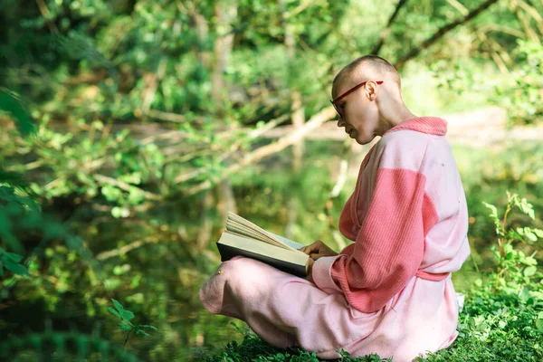 A woman with a short haircut reads a book on the shore of the lake against the backdrop of green nature. Sunny day. A woman in a bathrobe.