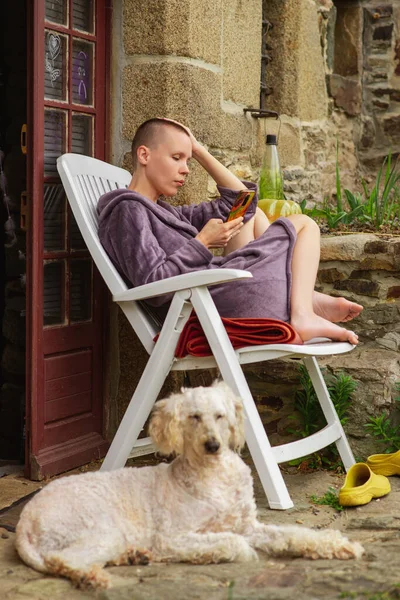 A bald woman is resting, sitting on a chair looking at the phone, a dog lies nearby. Rest on the porch of the house.