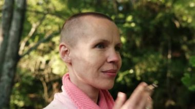 Kel kadın dışarıda sigara içiyor. Yeşil bitkilerin arka planında sabahlık giymiş kısa saçlı bir kadın..