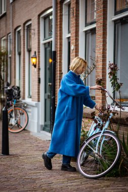 Hollanda 'da uzun mavi paltolu bir kadın. Rotterdam 'da seyahat ederken bir turist. Yetişkin bir kadın bisiklete biner.