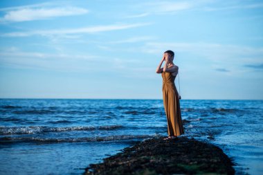 Bir kadın okyanusta batan güneşe dua eder. Uzun elbiseli, kısa saçlı orta yaşlı bir kadın..