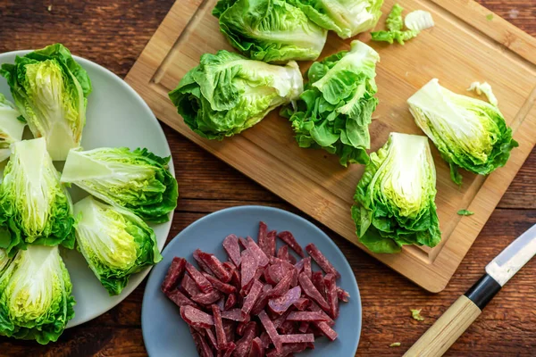 Fresh green lettuce cut into halves on a wooden cutting board. View from above. Chopped sausage. Recipe for stewed salad with meat. View from above
