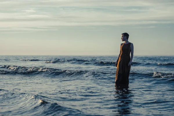 stock image A woman is standing in a long dress in the water in the ocean or sea. A woman with short hair looks at the sunset. Reflections, crisis.