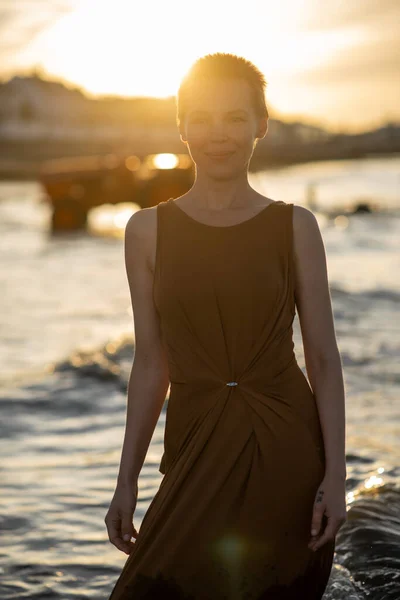 A woman with a short haircut in the rays of the sunset stands against the background of the water of the ocean. Smiling expression.