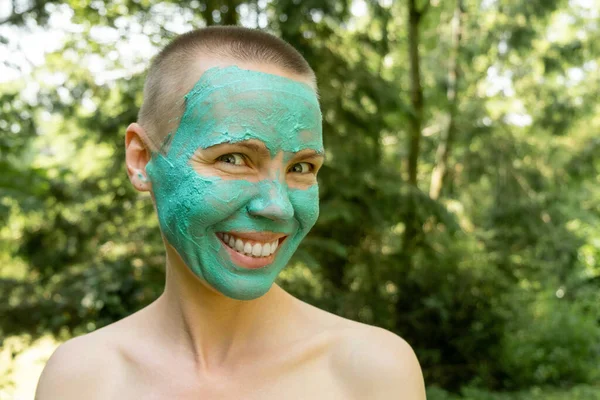 A middle-aged woman in a cosmetic mask smiles broadly and looks at the camera against the backdrop of a green forest. Woman with short hair.