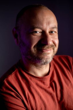 Yakın çekim, orta yaşlı, tıraşsız, sıradan görünümlü bir adamın samimi ifadesini ve yaklaşılabilir davranışını yakalıyor.