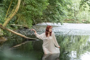 Beyaz elbiseli genç bir kız, yeşil yapraklı bir arka planda gölde duruyor. İvan 'ın yıkandığı gün.