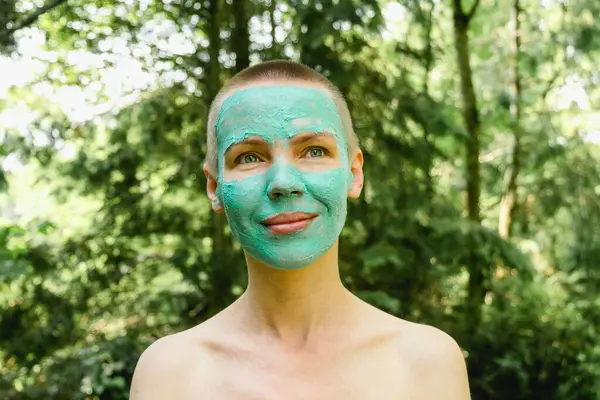 A middle-aged woman with a short haircut wearing a green cosmetic mask against a background of trees. Freshness, rejuvenation concept.