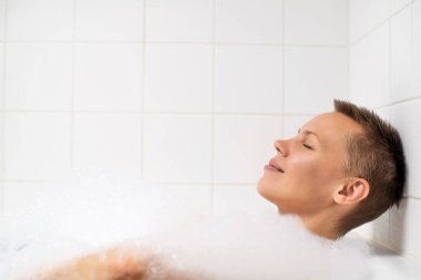 Kısa saçlı, köpüklü bir küvete dalmış bir kadın. Metin için yer