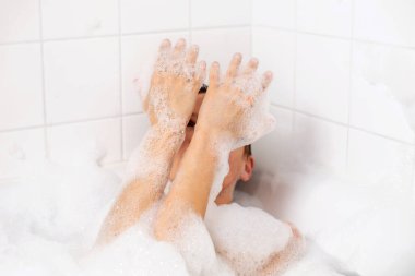 Bir kadın bol köpükle banyo yapar. Elleri köpükle kaplı, gülümsüyor ve elleriyle dışarı bakıyor.