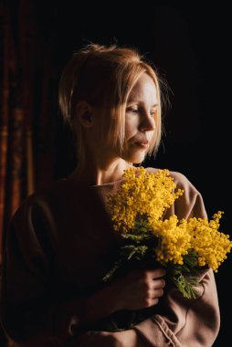 Karanlıkta, orta yaşlı bir kadın acı çekerek oturuyor, bir fener gibi gülümsüyor, elinde bir buket sarı çiçek, ışıldayan bir güzellik