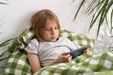 Yatakta uzanmış, 9-10 yaşındaki bir çocuk cep telefonuyla meşgul oluyor, çevrimiçi alemlerde geziniyor, oyunlara dalıyor, rahatlamayı sembolize ediyor ve hiçbir şey yapmama fikrini simgeliyor.