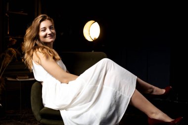 Karanlıkta oturan, dalgalı saçlı genç bir bayan sandalyeyi şereflendiriyor, gülümsemesi kameraya yöneltiliyor, odanın kararsızlığında sıcak bir kontrastlık yaratıyor.