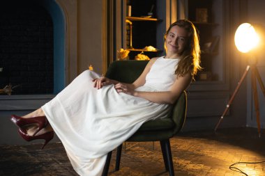 Karanlıkla çevrili, saçı dalgalı genç bir bayan koltuğundan kameraya sırıtarak odanın loş bir kontrastını oluşturuyor.