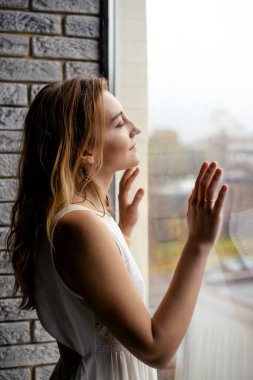 Beyaz zarafetle süslenmiş genç kadın, geniş pencerenin yanında duruyor. Yüzü sabah ışığıyla aydınlanıyor. Bu, güzel bir günün başlangıcını sembolize eden iyimserliğin bir resmi.