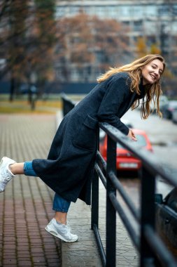 Beyaz spor ayakkabılı, uzun saçlı, korkuluklara yaslanan ve gülümseyen Mutlu Palto Günü kadını. Tatil zamanı, dinlenme