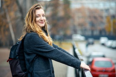 Uzun saçlı, sırtında sırt çantası olan gri paltolu bir kadın kameraya bakıyor ve sokak manzarasına gülümsüyor.