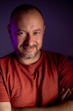 Orta yaşlı, tıraşsız, çekici bir yüz ifadesi olan, sıradan bir görünüm takınan ve hafif bir gülümseme takınan bir adamın yakın plan portresi.