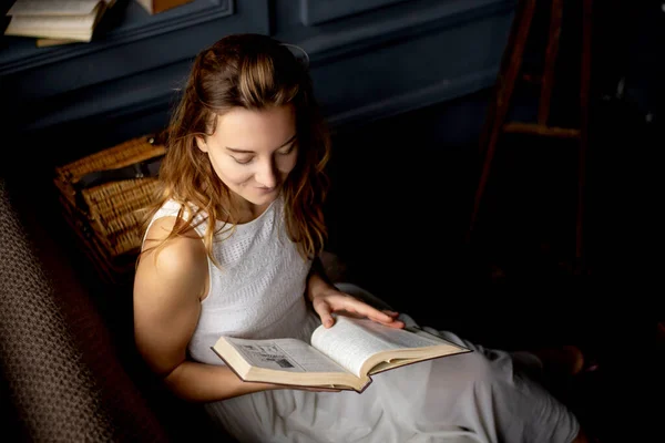 In the soft glow, a young woman with flowing hair smiles as she reads a book, seated on the sofa. The picture captures the essence of reading, reflecting her genuine passion for literature