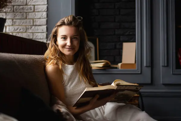 A young lady, her long locks framing her face, smiles while engrossed in a book, comfortably seated on the sofa. The image embodies the joy of reading and love for literature