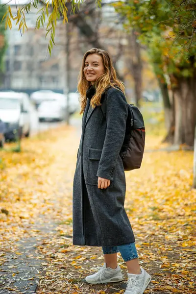 A young woman, her figure cloaked in a lengthy gray coat, carries a backpack, wandering through an autumnal city street carpeted with golden leaves, her smile reflecting the season's serenity