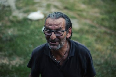 60 yaşındaki Sırp bir son sınıf öğrencisi, gözlüklü ve uzun saçlı, hareketli sohbetler ve bulaşıcı bir gülümsemeyle açık havada eğleniyor.