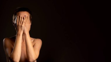 Siyah bir arkaplan önünde, orta yaşlarda kısa saçlı bir kadın elleriyle yüzünü gizler, olumsuz ve depresyon duygularını aktarır.