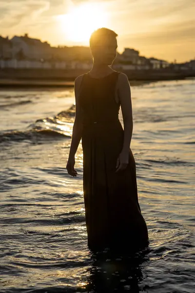 A woman with a short haircut in the rays of the sunset stands on the ocean in the water and smiles.