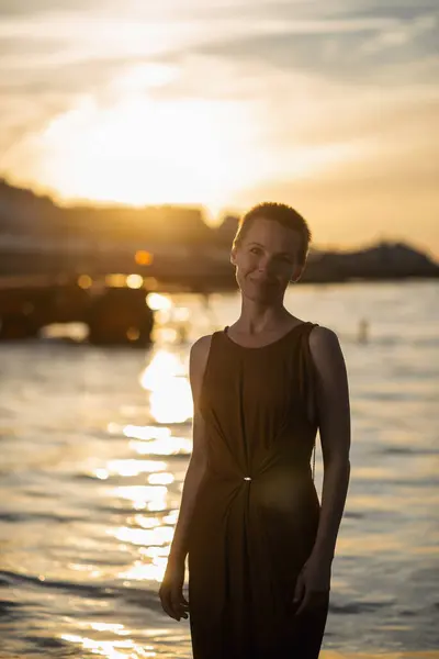 A woman with a short haircut in the rays of sunset stands in the ocean.