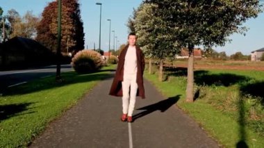 Orta yaşlı bir kadın şık bir ceket giyer, bu güneşli günde sakin bir yolda yol alır, özgüven ve zarafet saçar.